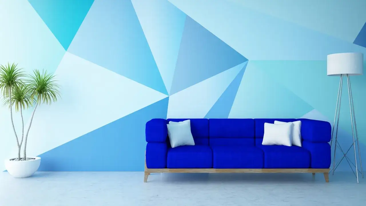 Niebieska sofa z białymi poduszkami na tle geometrycznych wzorów na ścianie. Obok stoi lampa podłogowa i doniczka z rośliną.