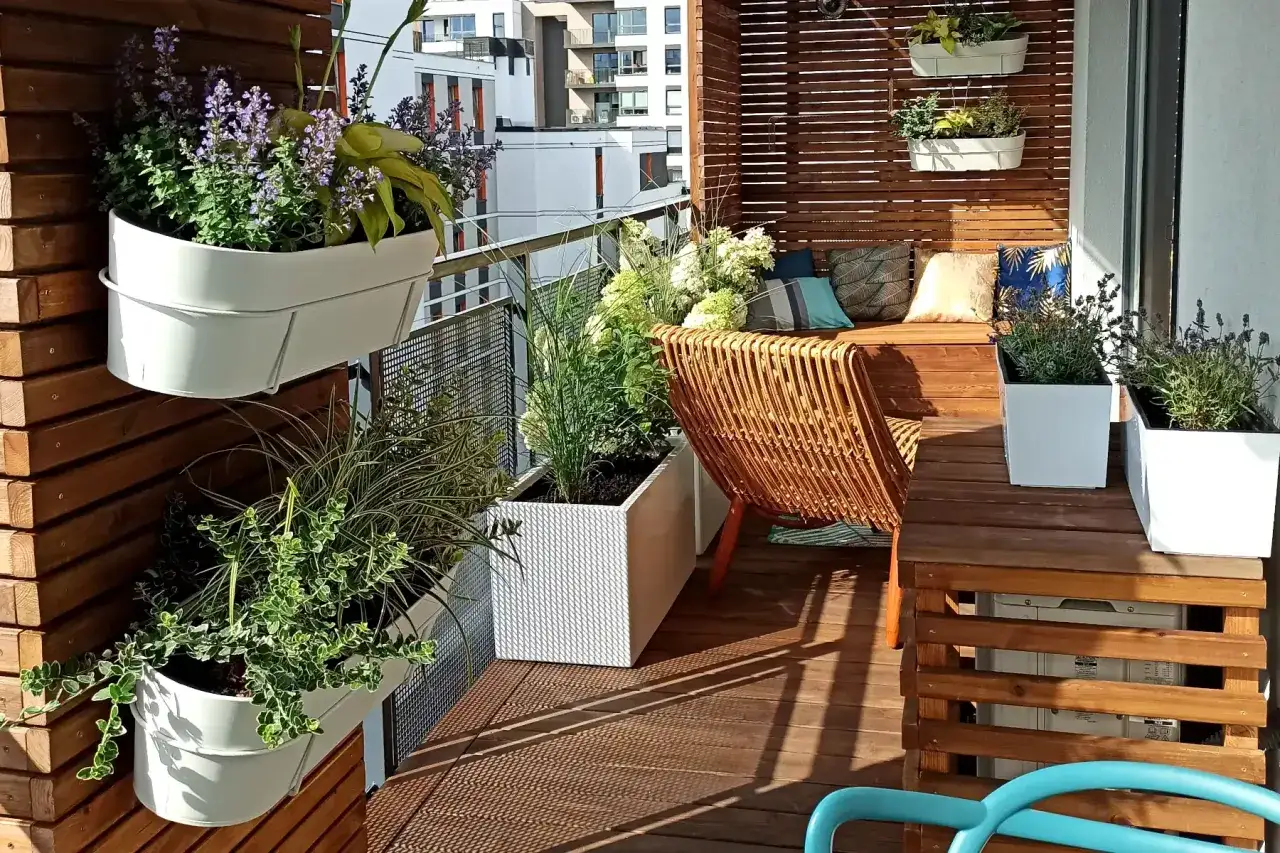 Zielony, kwitnący mały balkon w bloku. Wiszące donice z kwiatami, rattanowy fotel i drewniana ławka tworzą przytulne miejsce.